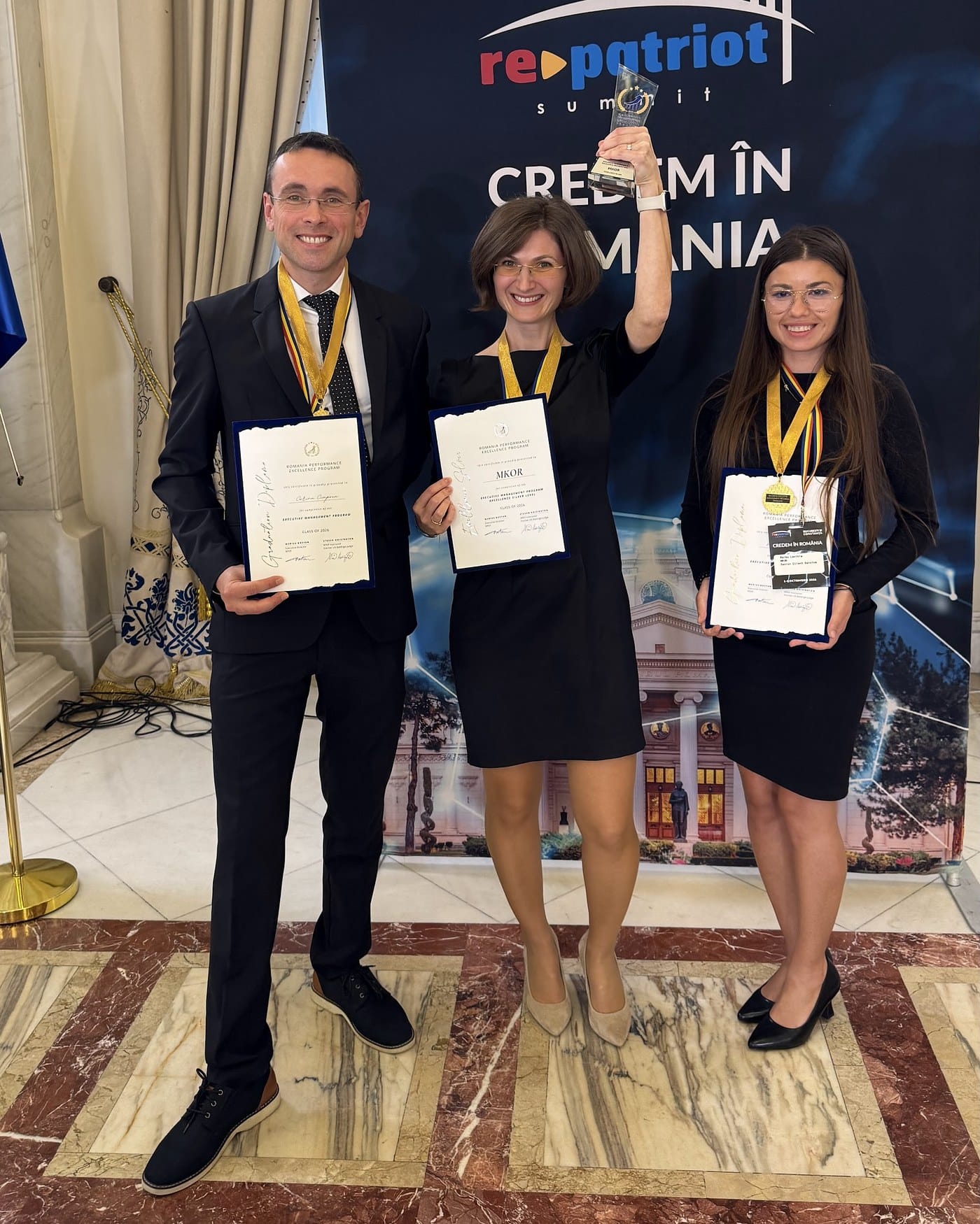 Trei membri ai echipei MKOR zâmbesc cu certificatele RPEP în mână, iar colega din mijloc ridică trofeul Excellence Silver, în fața panoului RePatriot Summit.