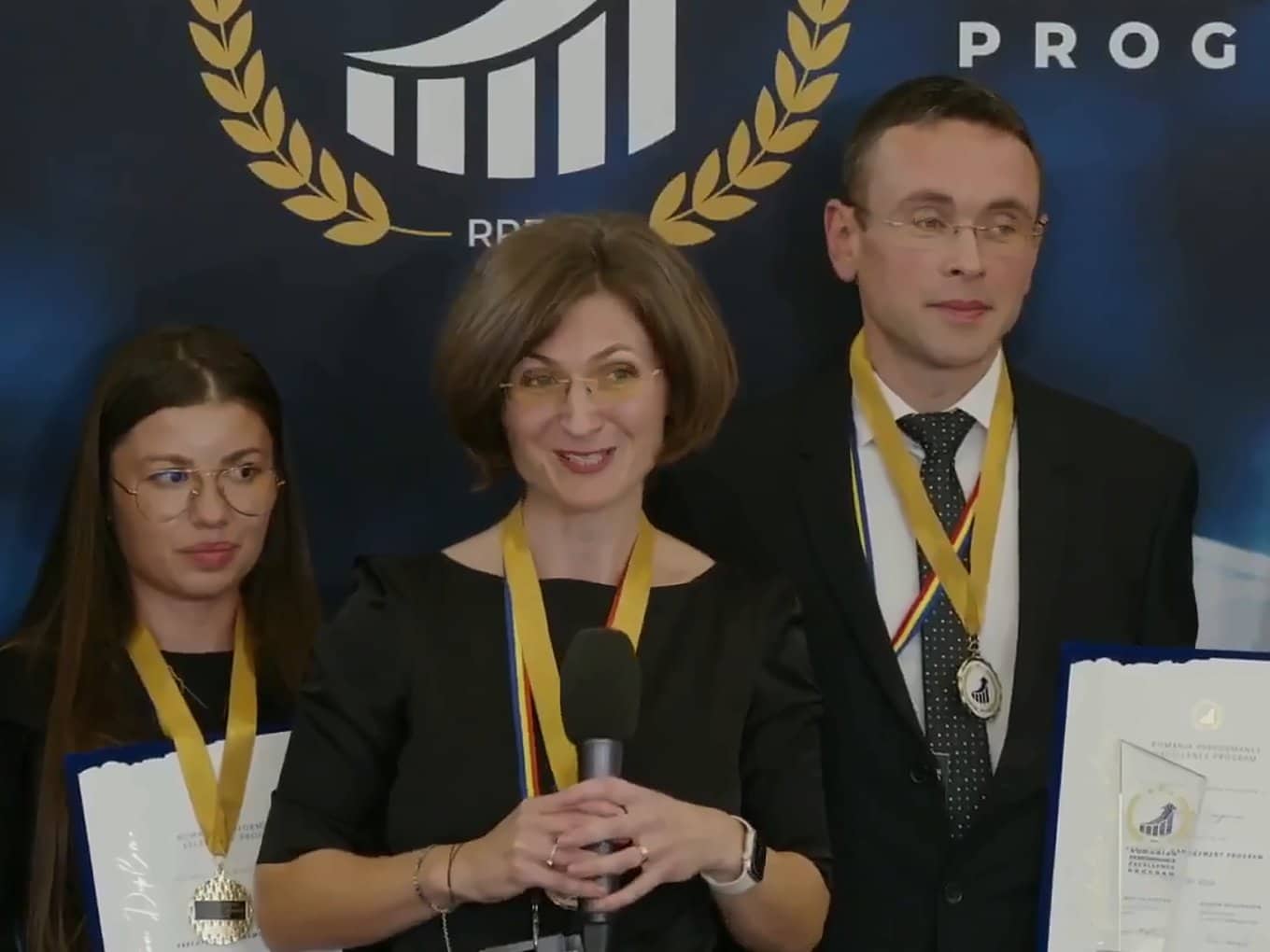 Trei membri ai echipei MKOR pe scenă, cu medalii și certificate, în fața panoului Romanian Performance Excellence Program; colega din mijloc vorbește la microfon.